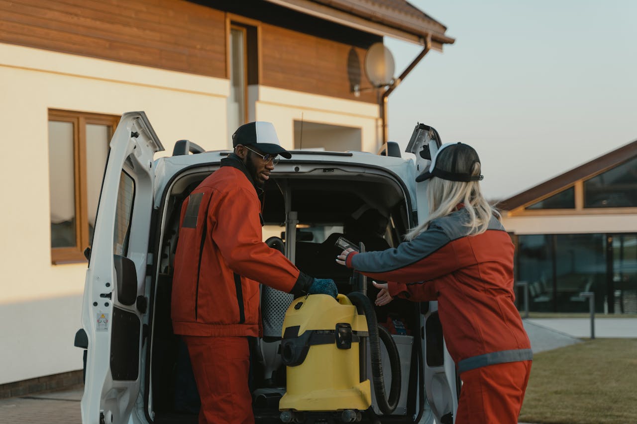 home-img Two cleaners in red uniforms load a vacuum into a white van outdoors.
