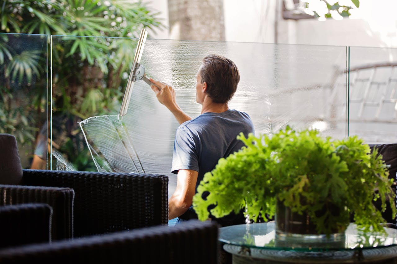 about-img-01 A man cleaning a glass barrier outdoors with a squeegee, surrounded by greenery.