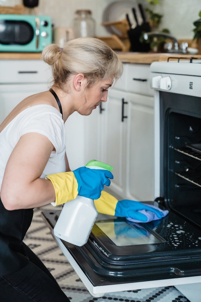 gallery-01 A woman uses a spray bottle and cloth to clean an oven in a modern kitchen setting.